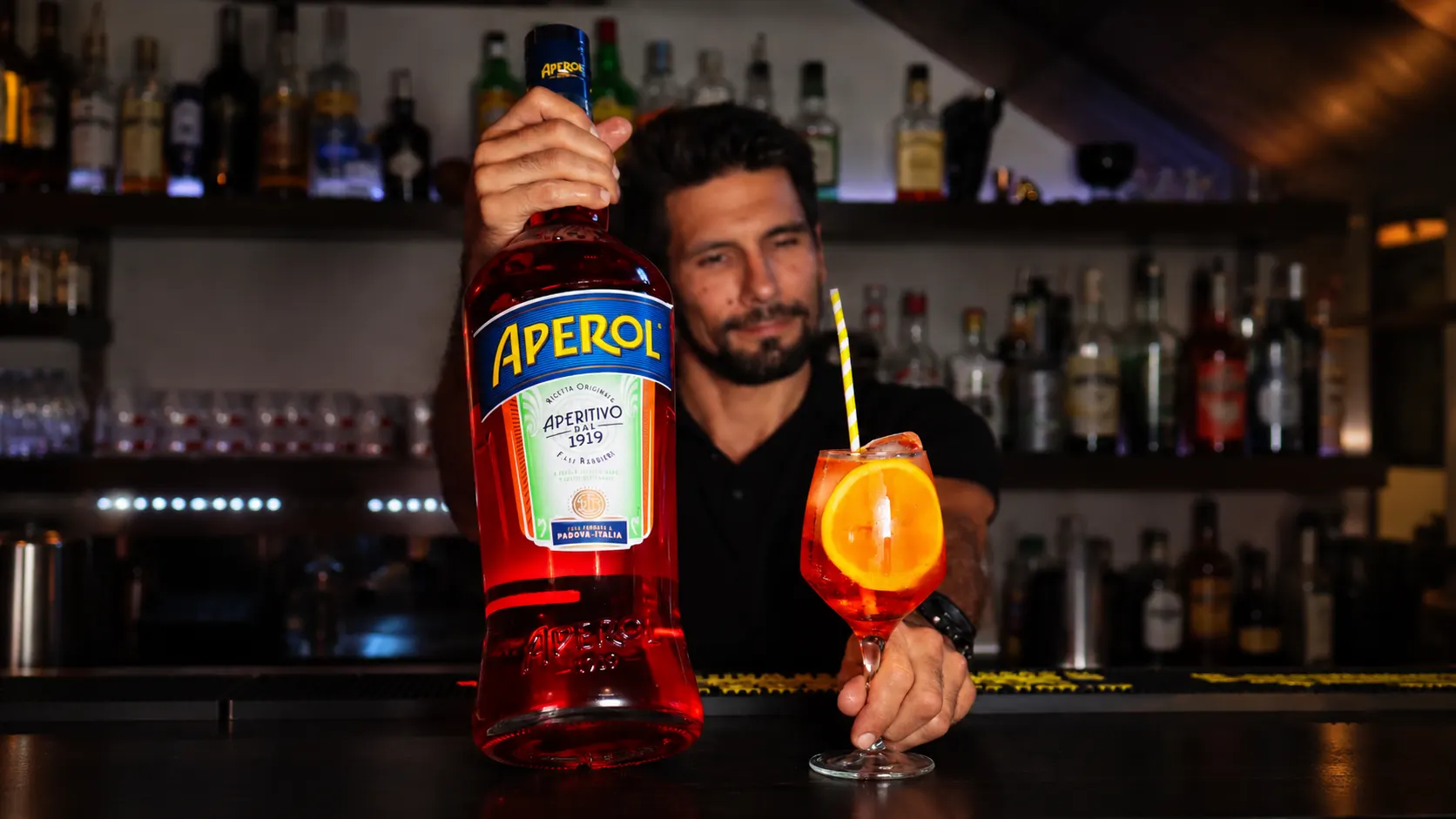 Um Aperol Spritz com rodela de laranja e palhinha, ao lado de uma garrafa grande de Aperol, no balcão.