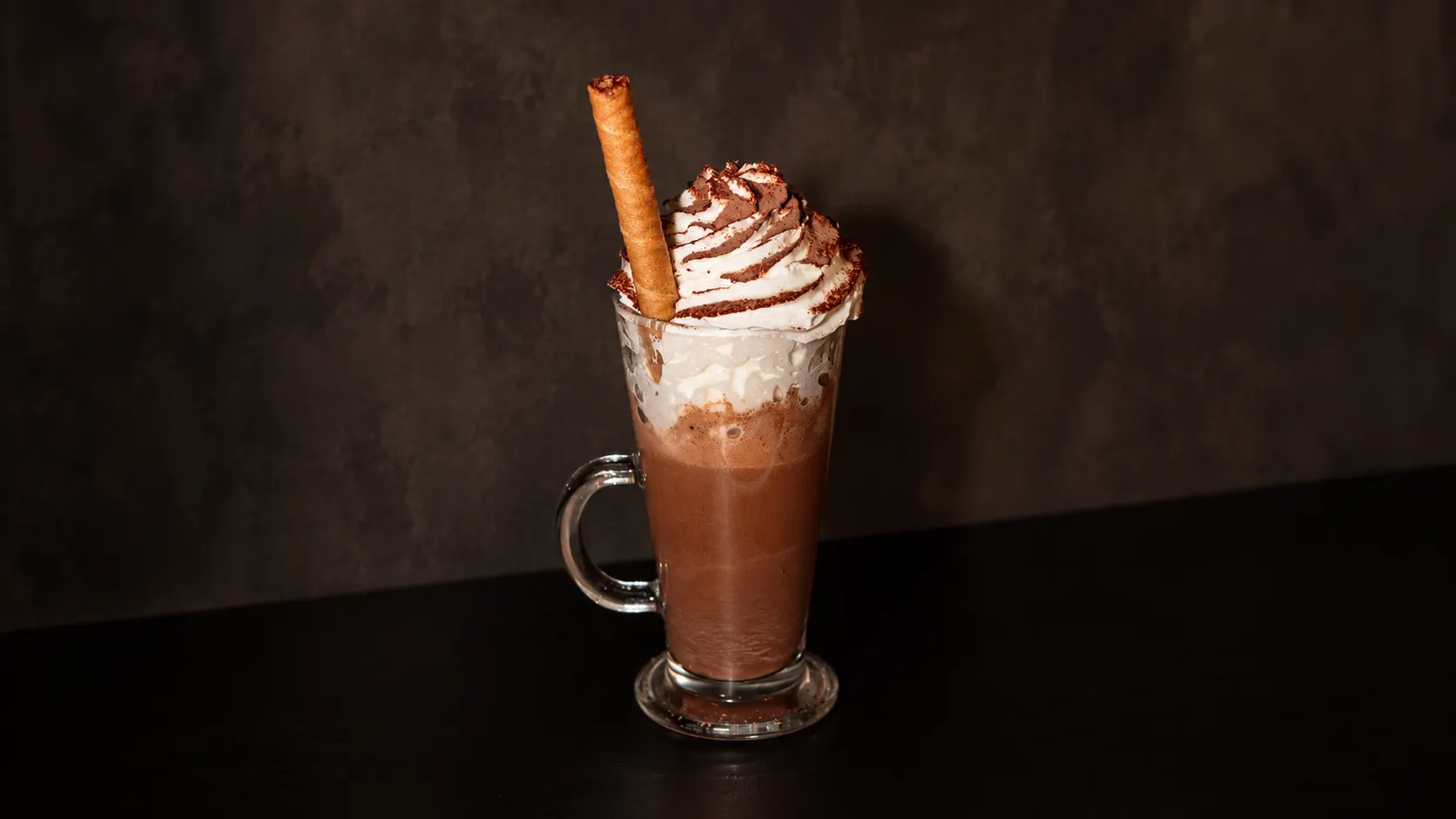 Um chocolate quente numa caneca de vidro, com chantilly, cacau e um barquilho.