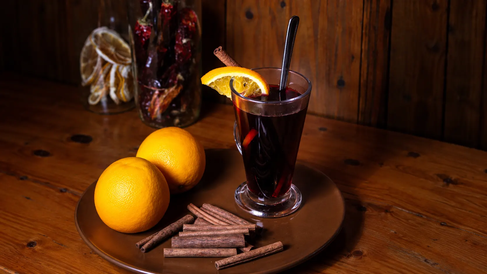 Um Glühwein, vinho tinto quente, numa caneca de vidro com uma rodela de laranja, ao lado de laranjas e paus de canela.