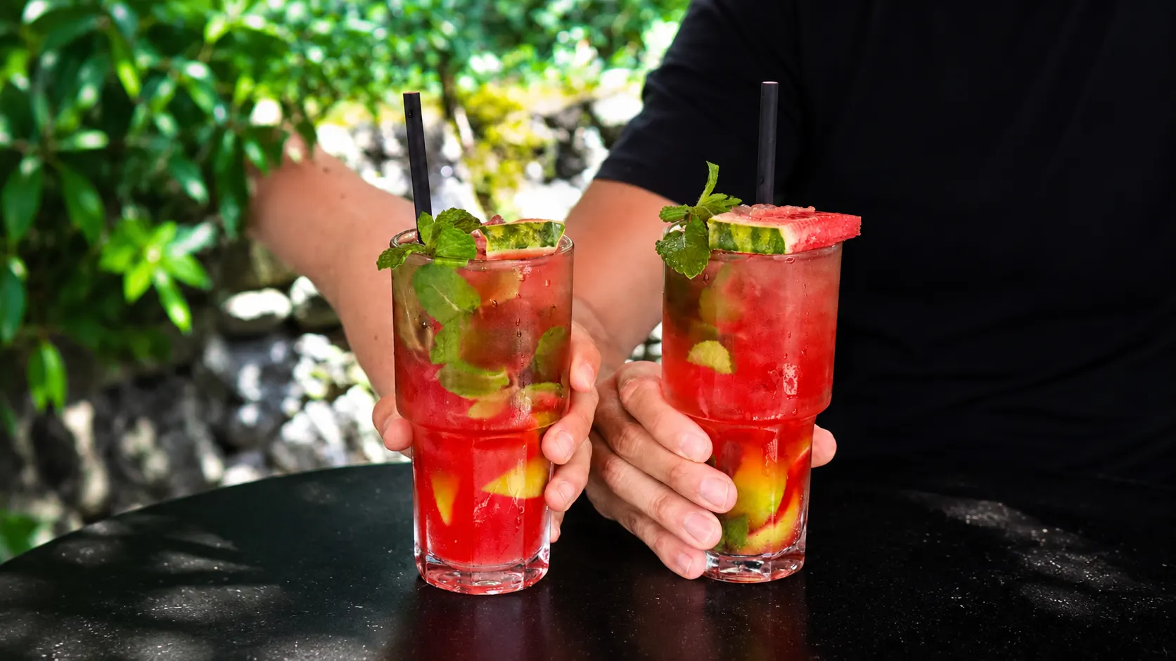 Dois mojitos de melancia, vermelhos, com hortelã e uma fatia de melancia, seguros por duas mãos.