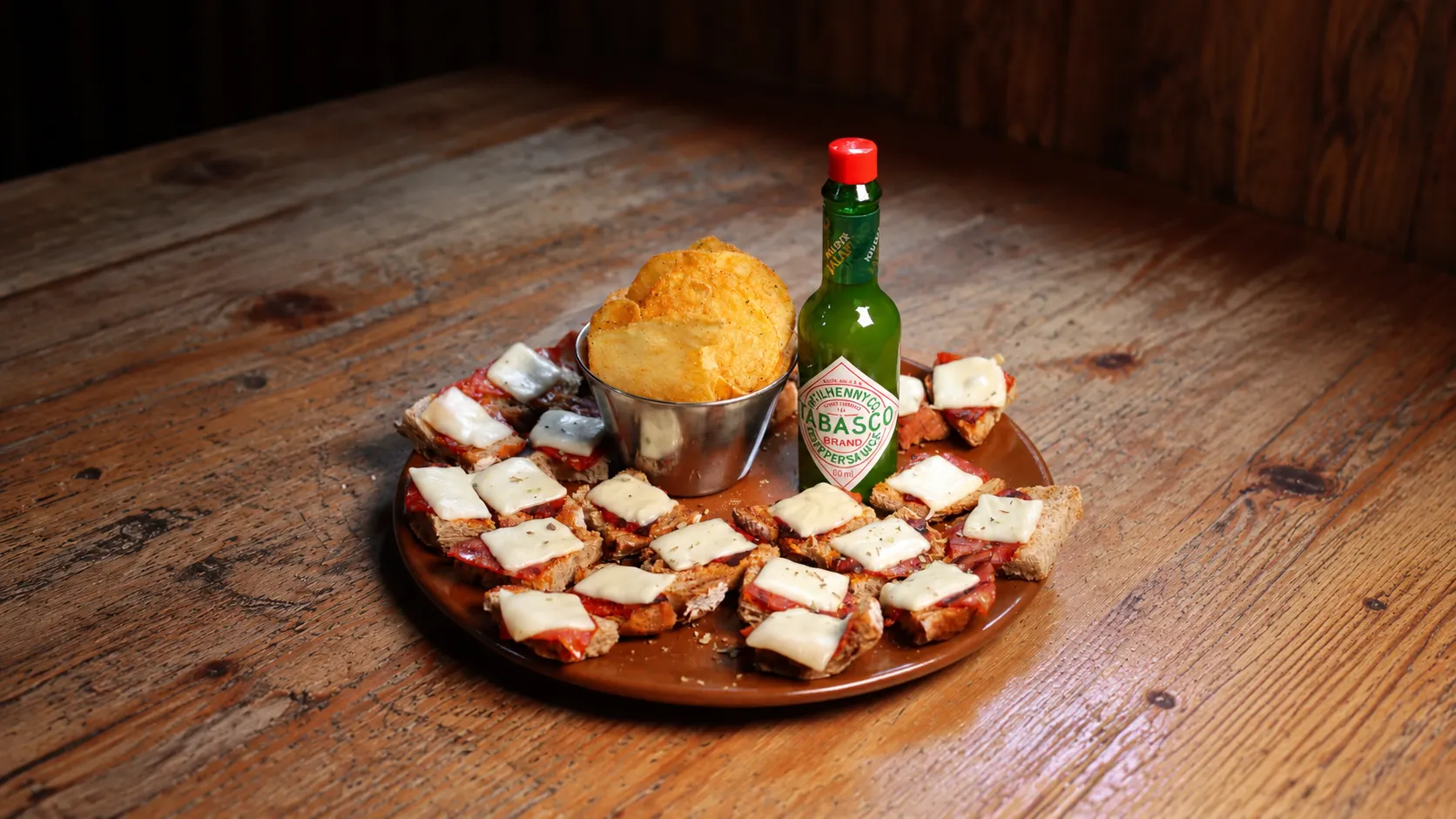 Uma travessa de tostinhas com chouriço e queijo à volta de uma taça de batata frita, com um frasco de tabasco.