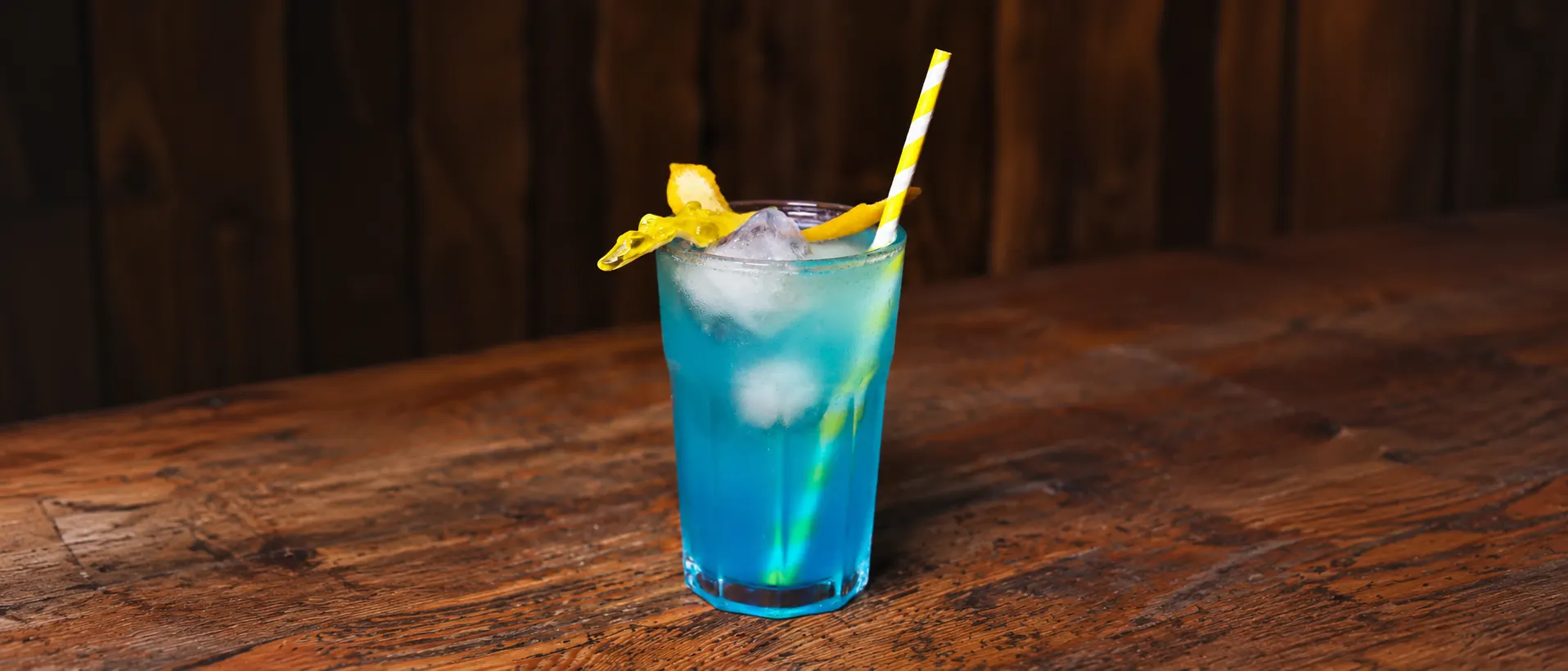 Um Unicórnio azul num copo de vidro com gelo, um tubarão de goma amarelo, casca de limão e uma palhinha às riscas amarelas, sobre um balcão de madeira.