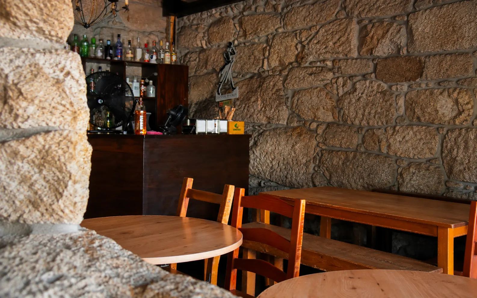 O canto do balcão com as garrafas, a parede de granito e mesas e cadeiras de madeira à frente.