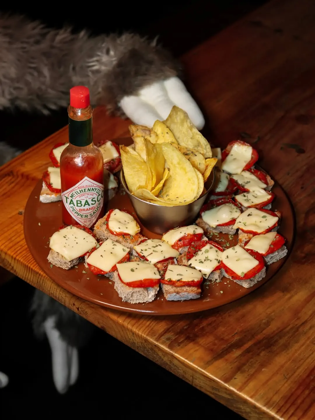 Uma travessa redonda com tostinhas de chouriço e queijo, uma taça de batata frita e um frasco de tabasco, sobre madeira.
